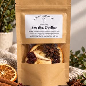May include: A brown kraft paper pouch containing a simmer pot kit labeled Lillard Mercantile Sweater Weather. The clear window reveals dried apple slices, orange slices, cranberries, and star anise. The label lists ingredients and directions.