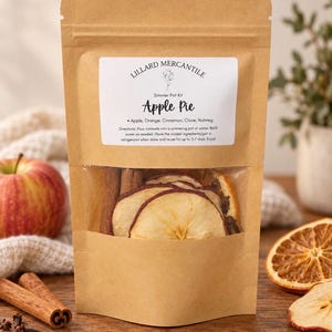 May include: A brown kraft paper pouch containing a simmer pot kit labeled Lillard Mercantile Apple Pie. The clear window reveals dried apple slices, cinnamon sticks, and cloves. The label lists ingredients and instructions for use.