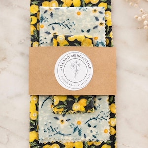 May include: A set of beeswax wraps featuring two distinct patterns: one with yellow lemons on a black background and another with white flowers on a light blue background. A brown paper band wraps around the center with the text Lillard Mercantile.