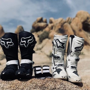May include: A pair of black and white motocross boots with a fox logo, and a pair of white and black motocross boots with a star logo. There are two pairs of tiny black and white baby booties.
