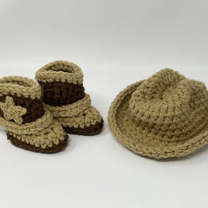 May include: A pair of crocheted baby booties and a matching hat in tan and dark brown yarn. The booties feature a decorative star on the side and a folded cuff design. The hat has a wide brim and a rounded crown, all crafted in a textured stitch.