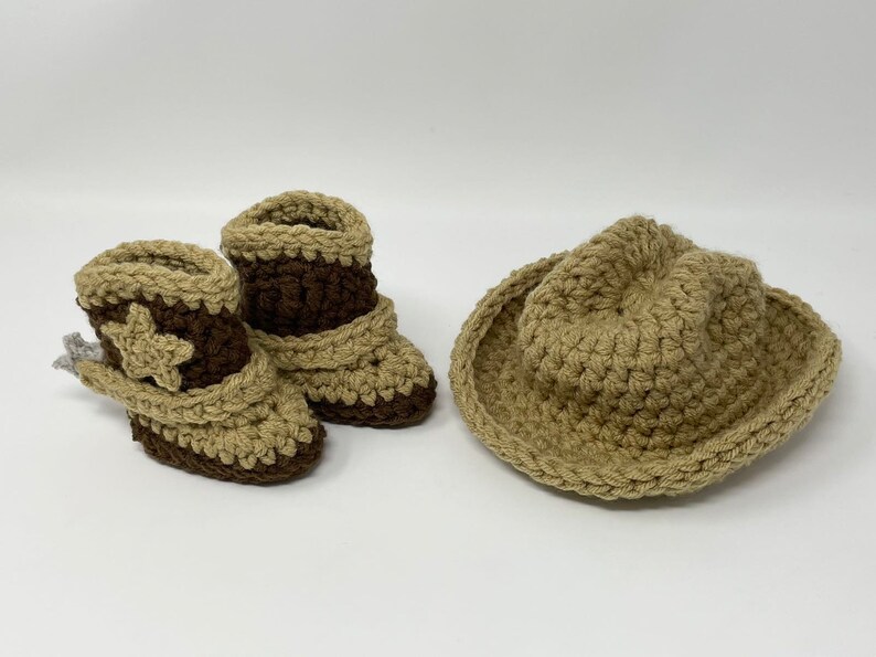 May include: A pair of crocheted baby booties and a matching hat in tan and dark brown yarn. The booties feature a decorative star on the side and a folded cuff design. The hat has a wide brim and a rounded crown, all crafted in a textured stitch.
