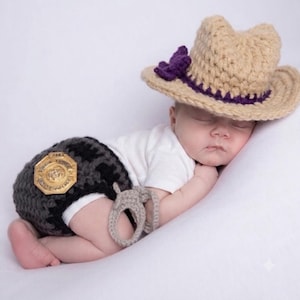 May include: A crochet set consisting of a tan cowboy hat with a purple flower accent, dark gray diaper cover shorts featuring a gold-colored police badge emblem, and a pair of light tan crochet handcuffs, worn over a plain white short-sleeved bodysuit.