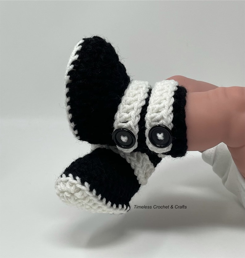 May include: Black and white crocheted baby booties with white trim and black buttons. The booties have a heart-shaped design on the buttons. Timeless Crochet & Crafts.