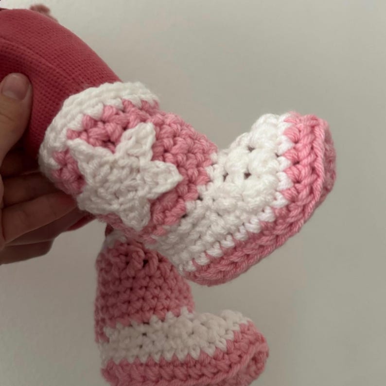 Puede incluir: Un par de patucos de beb&eacute; tejidos a ganchillo a mano con estilo de zapatillas altas. Los patucos est&aacute;n hechos de hilo rosa claro y blanco, con una prominente aplicaci&oacute;n de estrella blanca en el lateral del tobillo y cordones blancos.