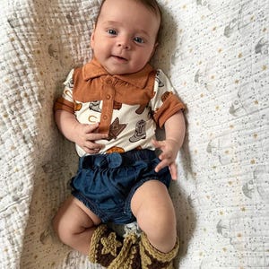 May include: A baby wearing a short-sleeved polo shirt with a cream background and a brown cowboy boot print, featuring a solid brown collar and placket. The outfit includes dark blue denim bloomers and crocheted tan and brown booties.