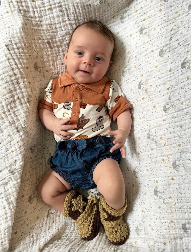 May include: A baby wearing a short-sleeved polo shirt with a cream background and a brown cowboy boot print, featuring a solid brown collar and placket. The outfit includes dark blue denim bloomers and crocheted tan and brown booties.