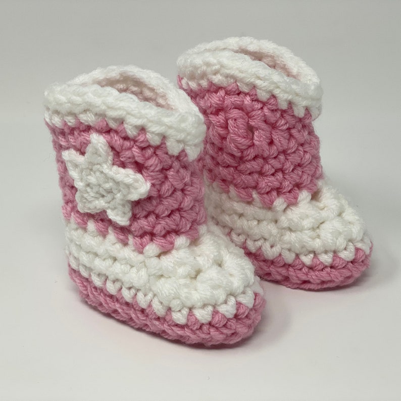 Puede incluir: Un par de patucos tejidos a mano con un dise&ntilde;o bicolor en hilo rosa suave y blanco. Los patucos tienen un pu&ntilde;o blanco, una puntera blanca y un cuerpo rosa con una prominente estrella tejida en blanco aplicada en el lado exterior.