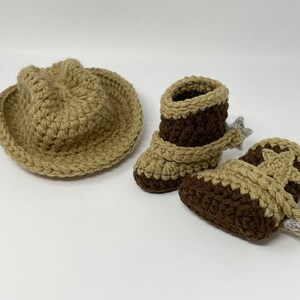 May include: A tan crocheted cowboy hat and a pair of brown and tan crocheted boots. The boots feature a tan strap across the ankle, a tan star detail on the side, and a small grey crocheted spur attached to the heel of each boot.