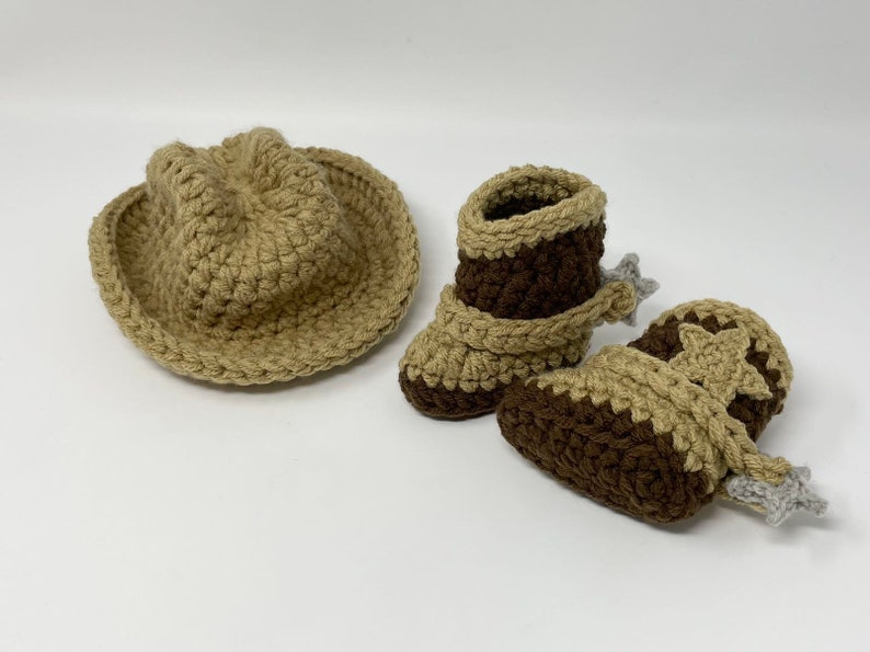 May include: A tan crocheted cowboy hat and a pair of brown and tan crocheted boots. The boots feature a tan strap across the ankle, a tan star detail on the side, and a small grey crocheted spur attached to the heel of each boot.