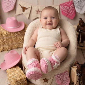 Puede incluir: Un beb&eacute; con un pelele de punto color crema y patucos de ganchillo rosas y blancos con estrellas. El beb&eacute; est&aacute; sentado en una silla mullida de color crema, rodeado de sombreros de vaquero rosas, balas de heno y sillas de montar en miniatura.