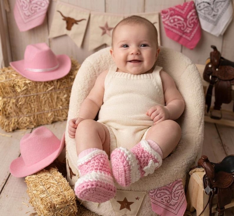 Puede incluir: Un beb&eacute; con un pelele de punto color crema y patucos de ganchillo rosas y blancos con estrellas. El beb&eacute; est&aacute; sentado en una silla mullida de color crema, rodeado de sombreros de vaquero rosas, balas de heno y sillas de montar en miniatura.