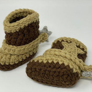 May include: A pair of handmade crochet baby booties designed to resemble cowboy boots. The booties feature a dark brown base with tan accents around the top and foot, finished with a small grey star-shaped spur attached to the heel area.