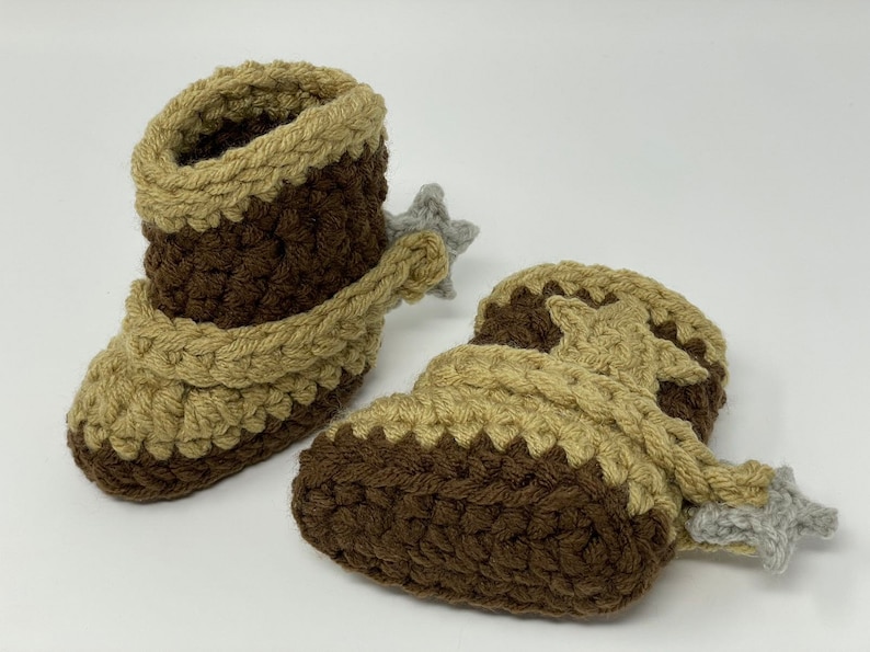 May include: A pair of handmade crochet baby booties designed to resemble cowboy boots. The booties feature a dark brown base with tan accents around the top and foot, finished with a small grey star-shaped spur attached to the heel area.