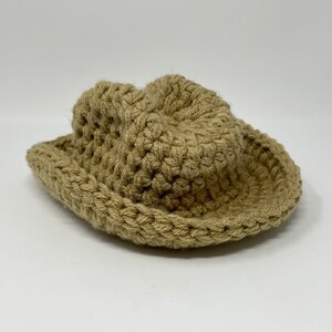 May include: A tan-colored, crocheted hat with a wide, curved brim and a rounded crown. The hat is constructed from thick, textured yarn in a consistent neutral beige tone, featuring visible interlocking loops throughout the entire structure.