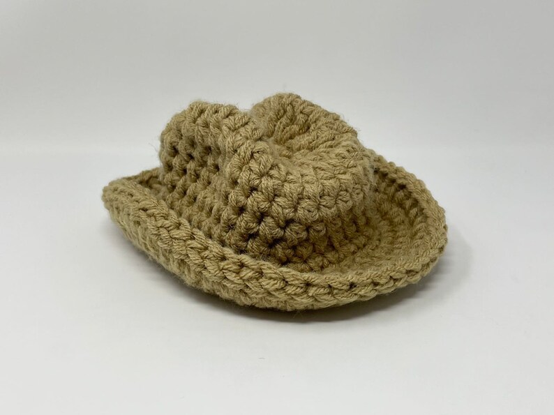 May include: A tan-colored, crocheted hat with a wide, curved brim and a rounded crown. The hat is constructed from thick, textured yarn in a consistent neutral beige tone, featuring visible interlocking loops throughout the entire structure.