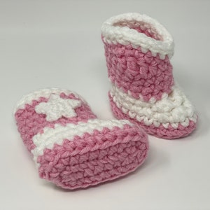 Puede incluir: Un par de patucos de ganchillo hechos a mano con un dise&ntilde;o bicolor en hilo rosa suave y blanco. Los patucos tienen un estilo de bota alta con una aplicaci&oacute;n de estrella blanca en el lateral y un patr&oacute;n de punto grueso y texturizado.