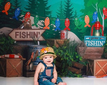May include: A child wearing blue denim overalls with a striped chest pocket and a tan crocheted hat with a small fish accent. The background features a fishing-themed display with wooden crates, a red lantern, and a banner of colorful fish.