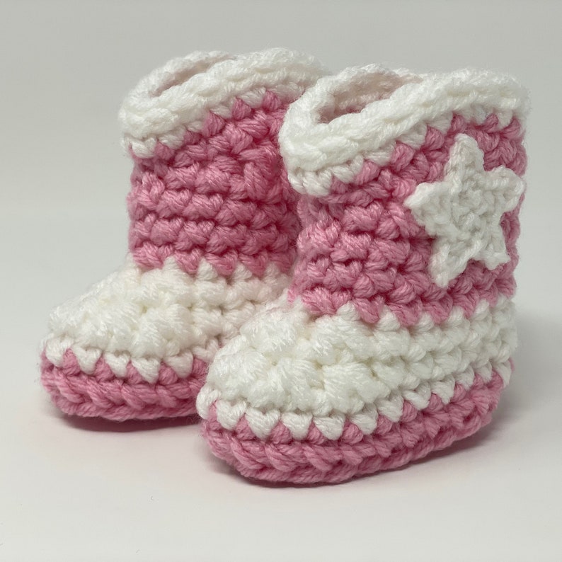 Puede incluir: Un par de botines tejidos a ganchillo a mano con un dise&ntilde;o bicolor en hilo rosa y blanco. Los botines tienen un patr&oacute;n de punto grueso y texturizado con una aplicaci&oacute;n de estrella blanca en el lateral y un pu&ntilde;o blanco en la abertura.