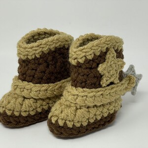 May include: A pair of handmade crochet booties designed to resemble cowboy boots. The footwear features a dark brown base and shaft with tan accents around the top, toe, and ankle strap. A tan star is attached to the side, and a grey spur is on the heel.