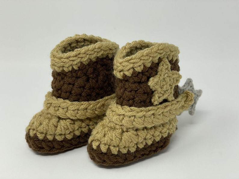 May include: A pair of handmade crochet booties designed to resemble cowboy boots. The footwear features a dark brown base and shaft with tan accents around the top, toe, and ankle strap. A tan star is attached to the side, and a grey spur is on the heel.