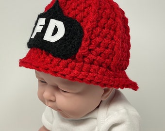 May include: Red crocheted firefighter hat with a black felt patch that says "FD" in white letters. The hat has a wide brim and is worn by a baby.