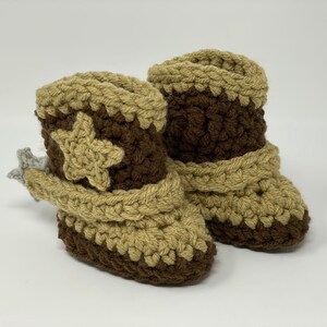 May include: A pair of crochet baby booties designed to look like cowboy boots. The boots feature a dark brown base and shaft with tan accents on the toe, top cuff, and a star applique on the side. A small grey crochet spur is attached to the heel.