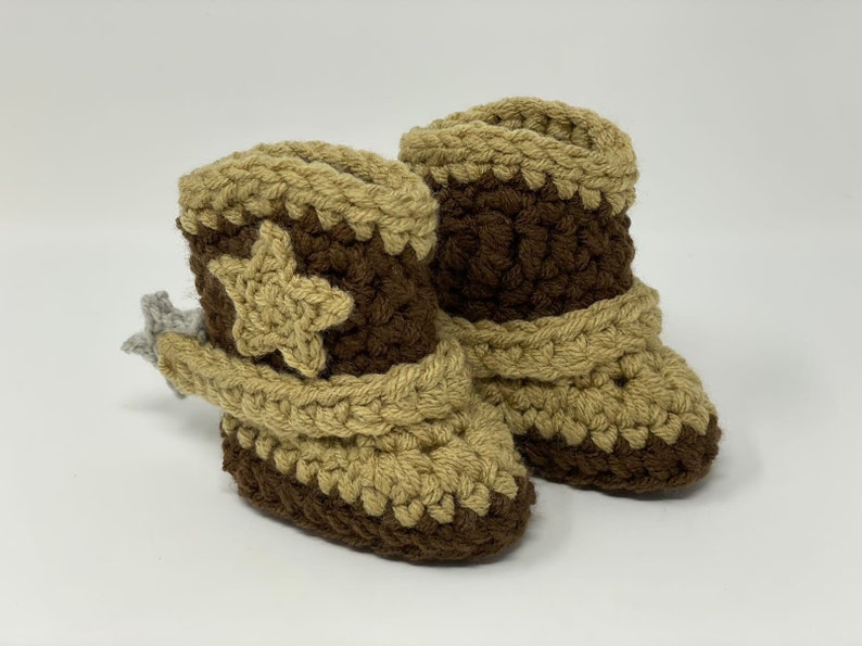 May include: A pair of crochet baby booties designed to look like cowboy boots. The boots feature a dark brown base and shaft with tan accents on the toe, top cuff, and a star applique on the side. A small grey crochet spur is attached to the heel.