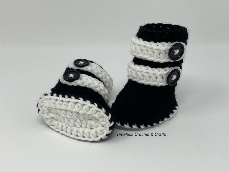 May include: A pair of black and white crocheted baby booties with black buttons. The booties have a white crocheted trim around the top and bottom.