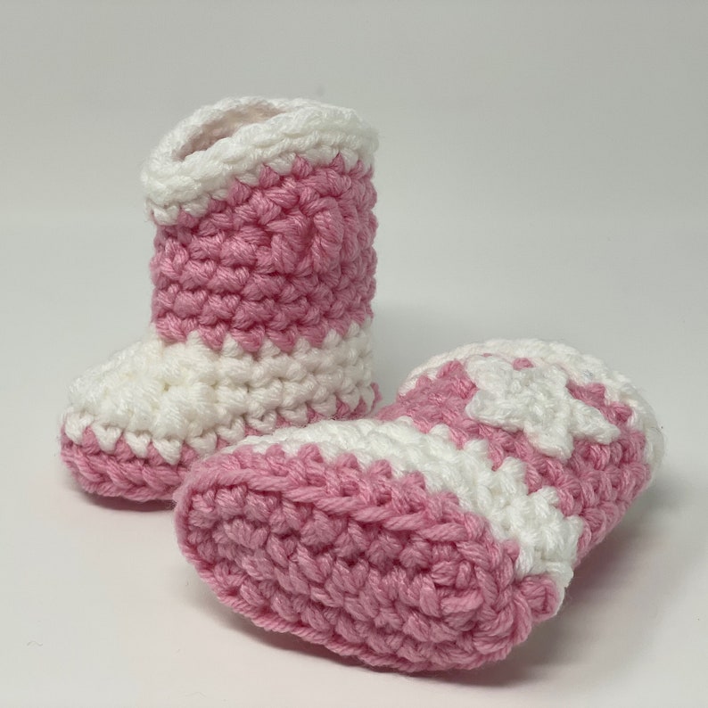 Puede incluir: Un par de patucos de ganchillo hechos a mano con un dise&ntilde;o bicolor de hileras alternas de hilo rosa suave y blanco. Los patucos tienen una estructura de ca&ntilde;a alta con un patr&oacute;n de punto grueso y texturizado y una puntera redondeada.
