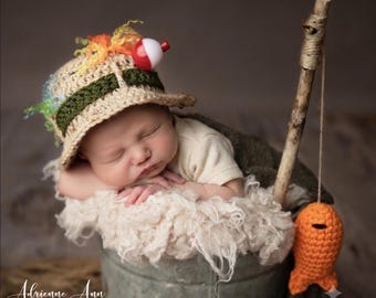 May include: A crocheted beige fishing hat with a green band, colorful yarn accents, and a red and white plastic bobber. The hat is worn by a sleeping infant resting in a metal bucket with a wooden fishing pole and a small orange crocheted fish.