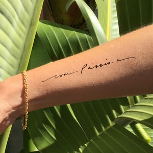 Puede incluir: Un tatuaje temporal en un brazo con las palabras "com. Passio." en una fuente cursiva delicada. Una pulsera dorada adorna la muñeca. El fondo presenta hojas verdes grandes, creando un entorno natural y vibrante.