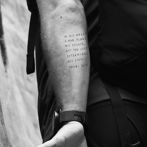 May include: A black and white photo of a person's arm with a tattoo that reads "In his heart a man plans his course, but the Lord determines his steps. Prov. 16:9".