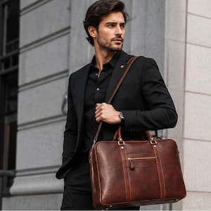May include: A dark brown leather briefcase featuring a rectangular shape, dual top handles, a detachable shoulder strap, and a front zippered pocket with gold-toned hardware. The bag is constructed with visible stitching and reinforced corners.