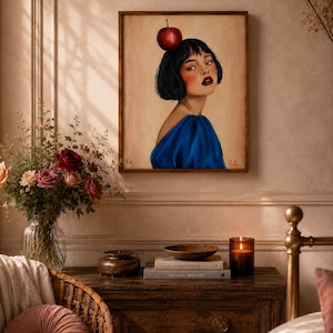 Peut inclure: Un portrait encadré d'une femme aux cheveux noirs et au rouge à lèvres rouge, vêtue d'une robe bleue et d'une pomme sur la tête. L'œuvre est exposée au-dessus d'une commode en bois avec une bougie, des livres et un bol décoratif.