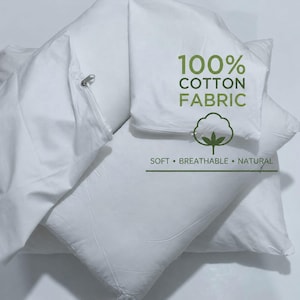 May include: A set of white pillowcases with zippers, featuring the printed text 100% COTTON FABRIC, a cotton plant icon, and the words SOFT • BREATHABLE • NATURAL. The fabric is plain white and smooth, showing a clear view of the zipper closure.