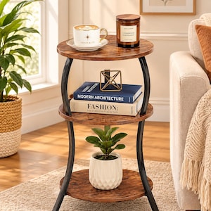 May include: A three-tier round side table with a dark wood grain finish and curved black metal legs. The top shelf holds a white coffee cup and a candle. The middle shelf displays two books titled Modern Architecture and Fashion.