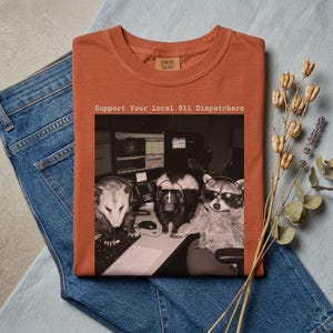 May include: A rust-colored t-shirt featuring a graphic of an opossum, a skunk, and a raccoon wearing headsets at a computer workstation. The text above the image reads, Support Your Local 911 Dispatchers. The shirt is laid flat on blue denim fabric.