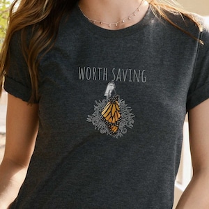 May include: A dark heather grey t-shirt with a graphic of a monarch butterfly emerging from its chrysalis. The text "WORTH SAVING" is printed above the graphic.