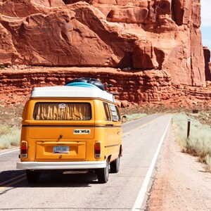 K&ouml;nnte beinhalten: Ein gelber Oldtimer-Van mit wei&szlig;em Dach f&auml;hrt eine W&uuml;stenstra&szlig;e entlang. Ein Surfbrett und Gep&auml;ck sind oben befestigt. Das Nummernschild lautet "EXPLORE". Rote Felsformationen und ein blauer Himmel im Hintergrund.