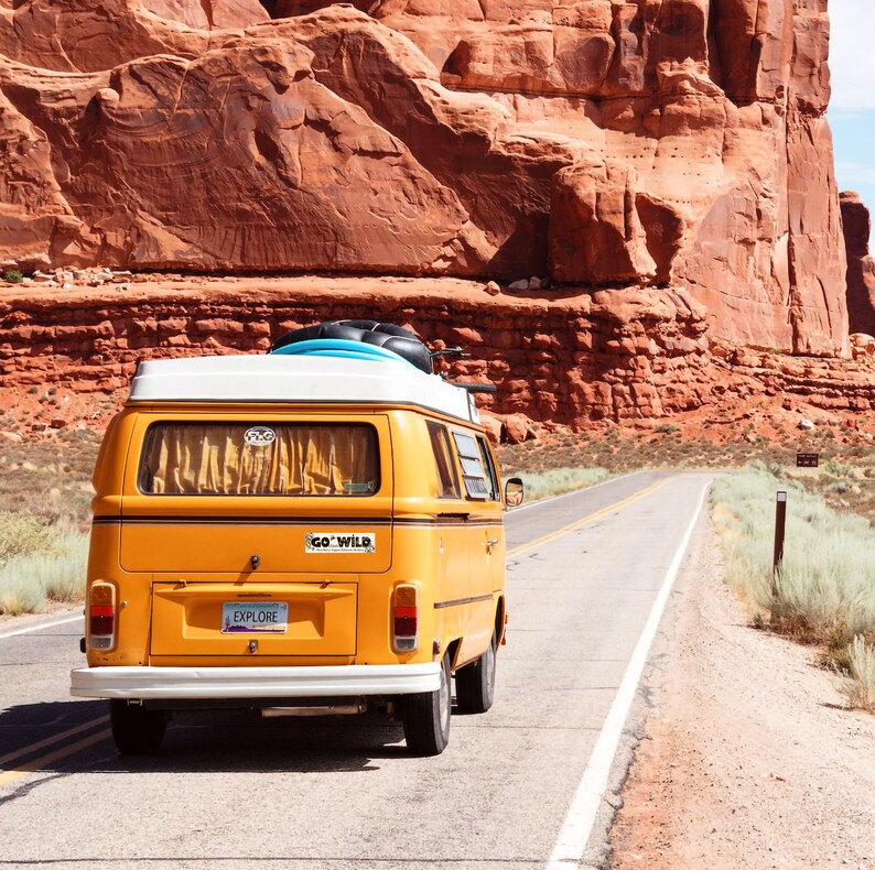 K&ouml;nnte beinhalten: Ein gelber Oldtimer-Van mit wei&szlig;em Dach f&auml;hrt eine W&uuml;stenstra&szlig;e entlang. Ein Surfbrett und Gep&auml;ck sind oben befestigt. Das Nummernschild lautet "EXPLORE". Rote Felsformationen und ein blauer Himmel im Hintergrund.