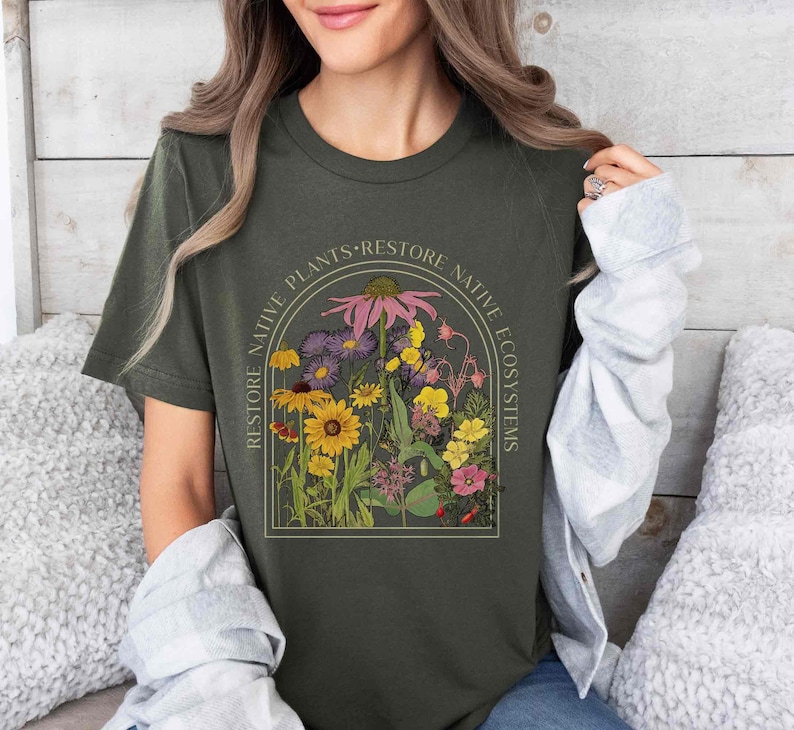 K&ouml;nnte beinhalten: Ein dunkelgr&uuml;nes T-Shirt mit einer Grafik einer Vielzahl von Blumen in einem kreisf&ouml;rmigen Rahmen. Der Text "RESTORE NATIVE PLANTS - RESTORE NATIVE ECOSYSTEMS" umgibt die Grafik.