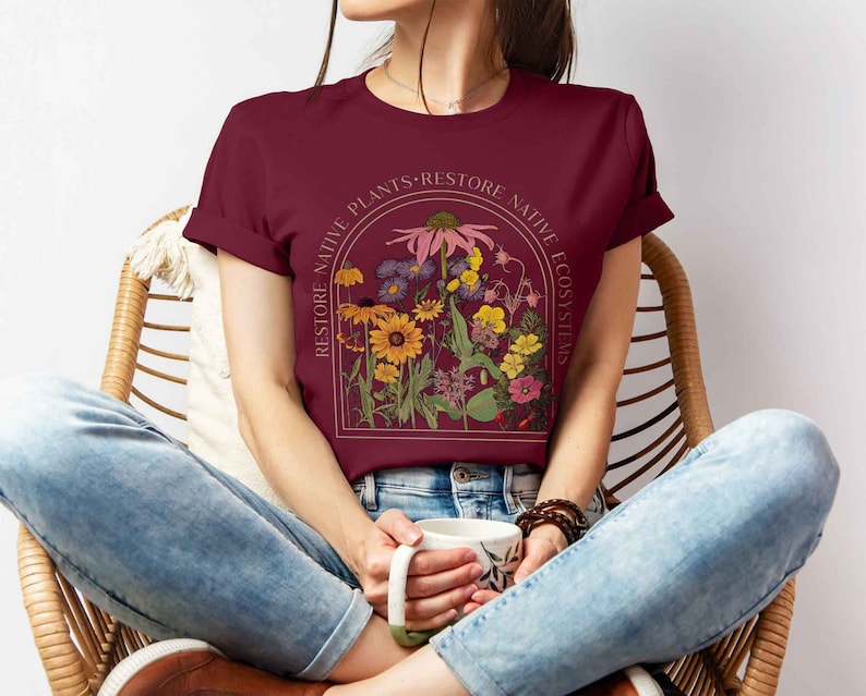 K&ouml;nnte beinhalten: Ein maroonfarbenes T-Shirt mit einem Grafikdruck von verschiedenen Blumen in einem Kreis mit dem Text "Restore Native Plants - Restore Native Ecosystems".