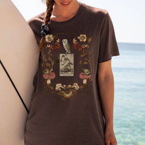 May include: A dark gray t-shirt with a vintage illustration of an owl, a bird, and butterflies surrounded by floral designs. The illustration is framed by a rectangle with the text "The Garden Sprite".