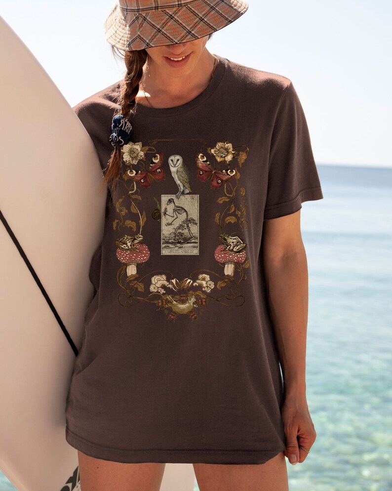 May include: A dark gray t-shirt with a vintage illustration of an owl, a bird, and butterflies surrounded by floral designs. The illustration is framed by a rectangle with the text "The Garden Sprite".