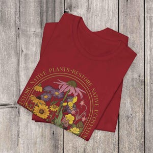 K&ouml;nnte beinhalten: Rotes T-Shirt mit einem goldenen Kreis-Motiv, das einen Strau&szlig; Wildblumen und den Text "Restore Native Plants - Restore Native Ecosystem" zeigt.