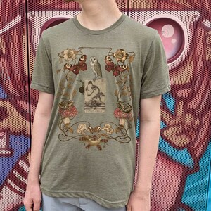 May include: A green t-shirt with a vintage illustration of a bird, butterflies, frogs, and mushrooms. The illustration is surrounded by a floral design.