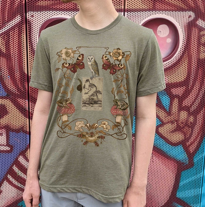 May include: A green t-shirt with a vintage illustration of a bird, butterflies, frogs, and mushrooms. The illustration is surrounded by a floral design.