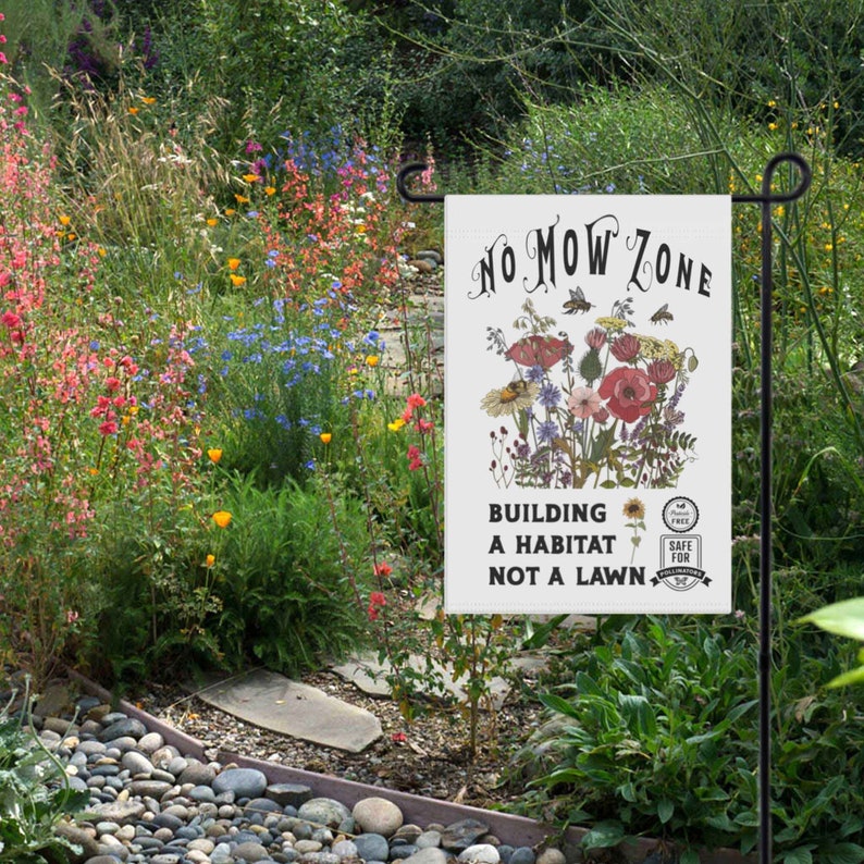 K&ouml;nnte beinhalten: Eine wei&szlig;e Gartenflagge mit dem Text "NO MOW ZONE" und einer floralen Illustration. Die Flagge lautet auch "BUILDING A HABITAT NOT A LAWN" und zeigt ein "SAFE FOR POLLINATORS"-Emblem. Die Flagge wird in einem Garten gezeigt.