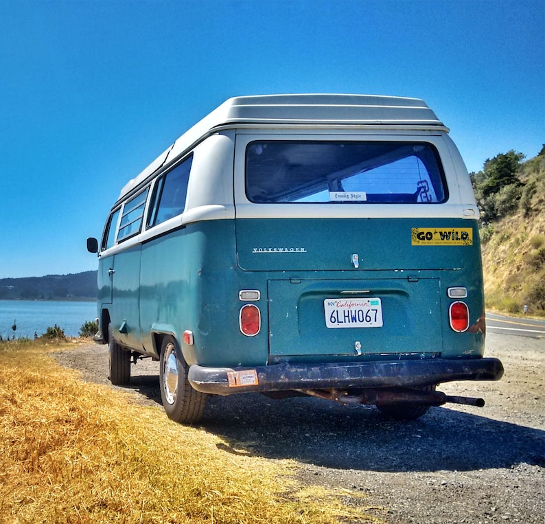 K&ouml;nnte beinhalten: Ein alter t&uuml;rkisfarbener und wei&szlig;er Volkswagen-Bus steht an einer Stra&szlig;e mit Blick auf einen See und H&uuml;gel. Der Bus hat ein kalifornisches Nummernschild und einen "GO WILD"-Aufkleber. Der Himmel ist strahlend blau.